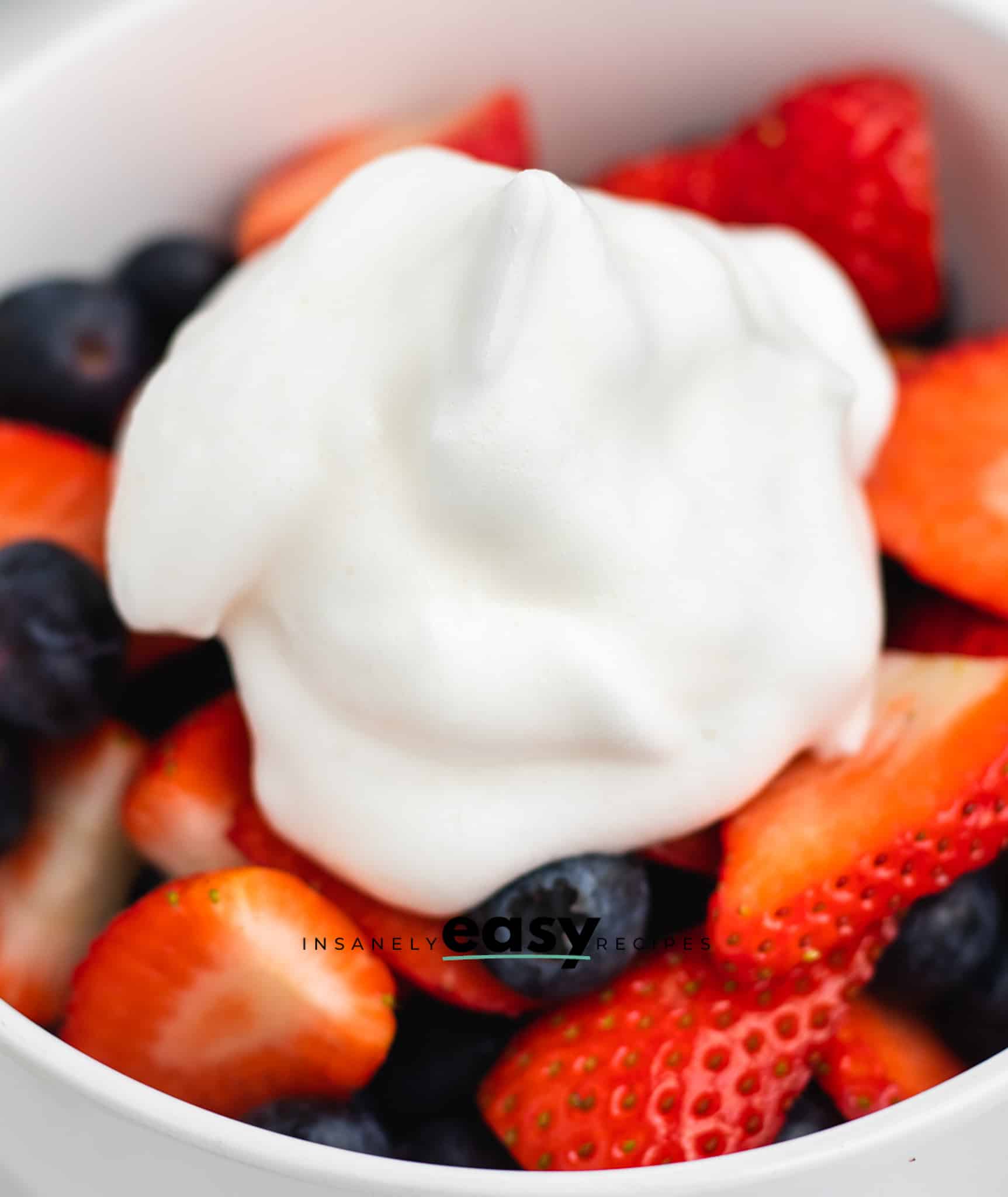 Closeup photo of a bowl of chopped berries, like blueberries and strawberries, topped with a dollop of aquafaba whipped cream.