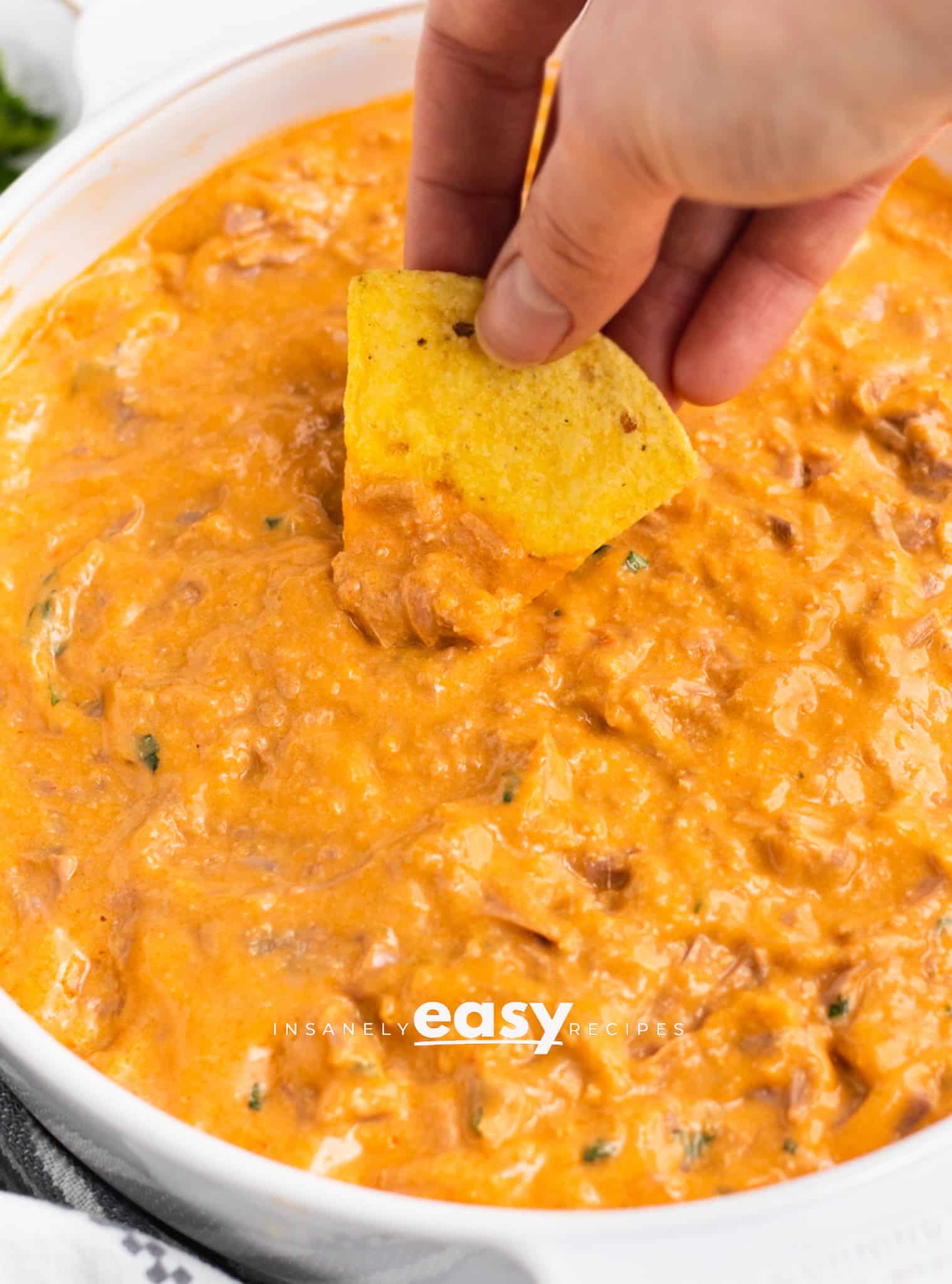 Photo of a hand dipping a corn tortilla chip into a shallow white dish, filled with vegan buffalo chicken dip. 