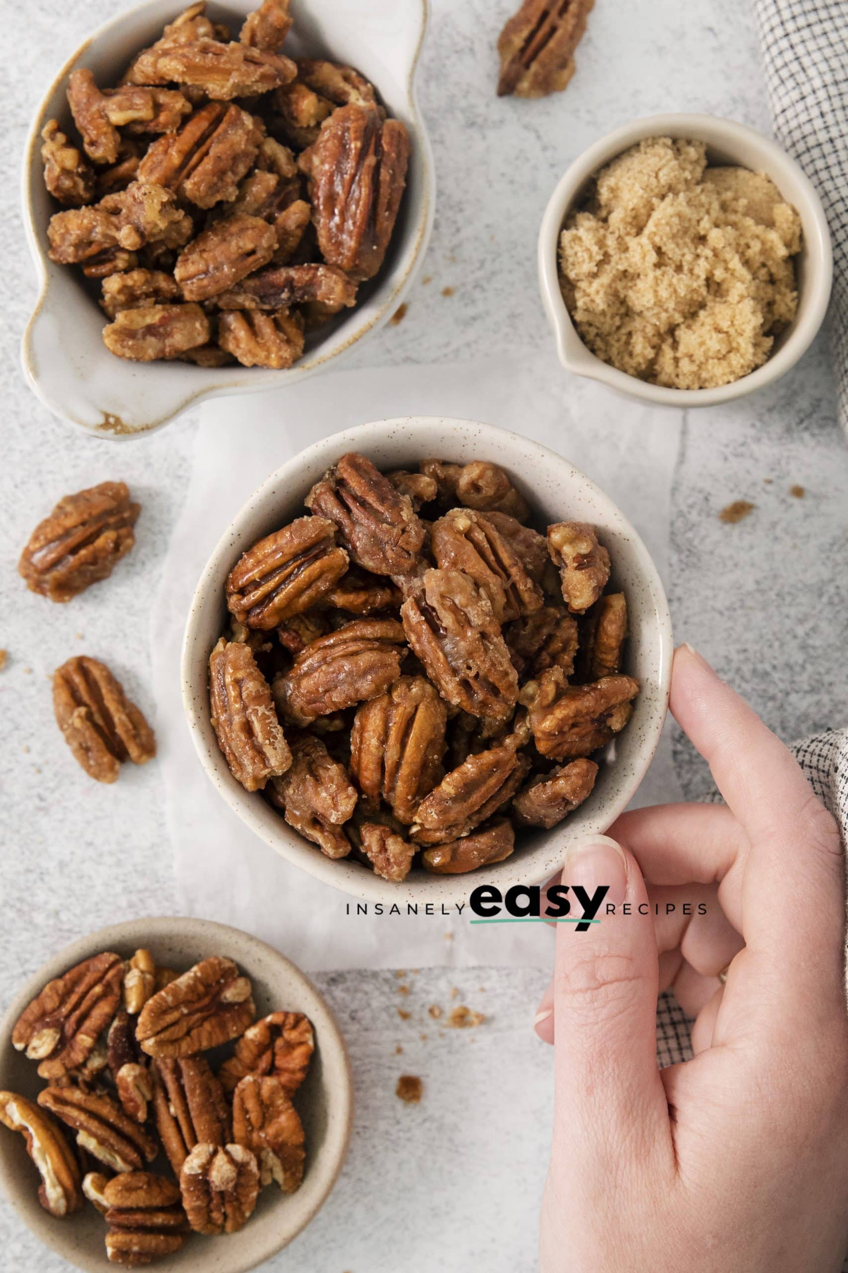 Top view photo of a bowl filled with Candied Pecans. There is a hand reaching in to grab the cup. The bowl is surrounded by other bowls, filled with brown sugar and raw pecans. 