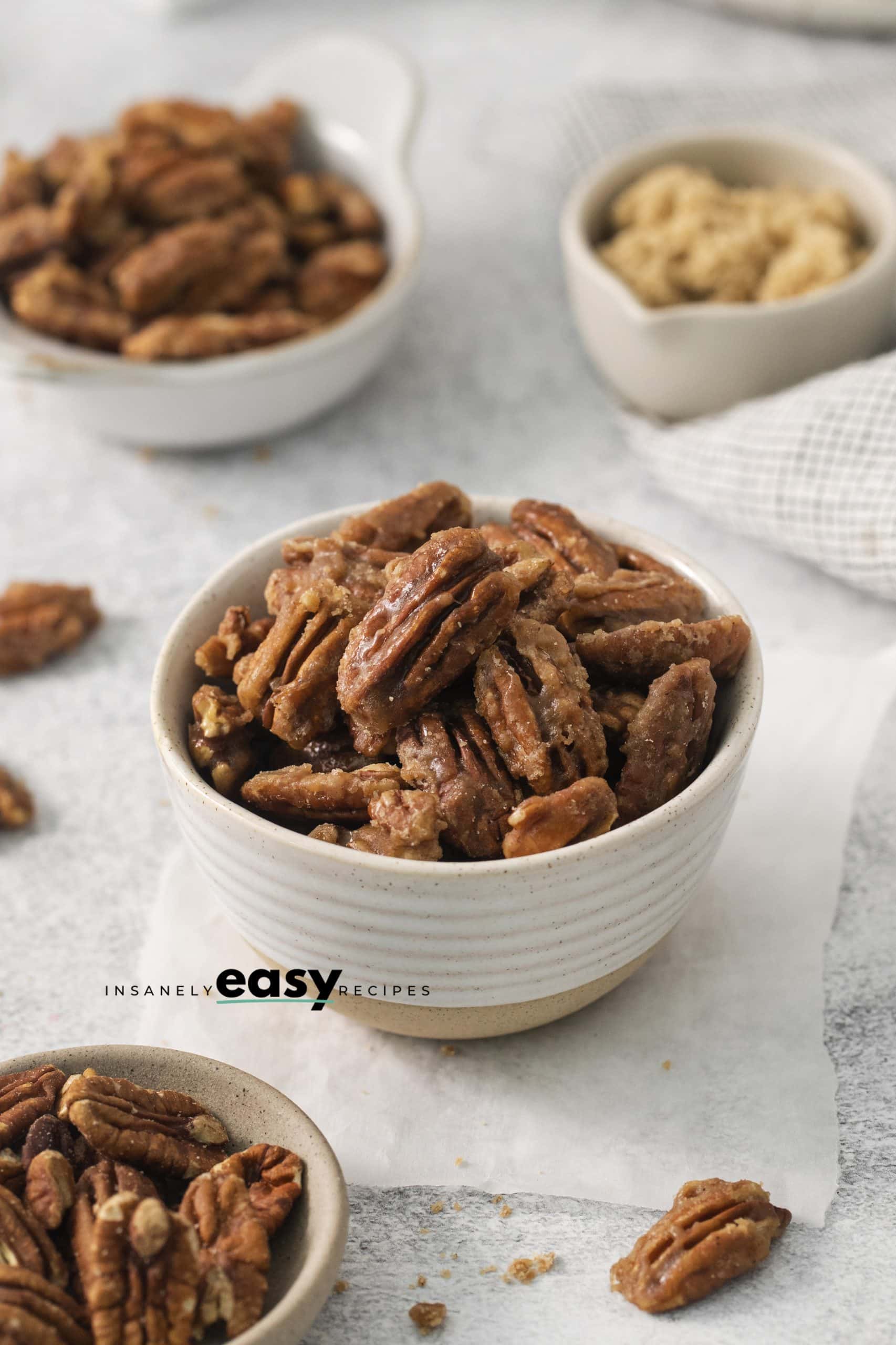 Top view photo of a bowl filled with Candied Pecans. The bowl is surrounded by other bowls, filled with brown sugar and raw pecans. 