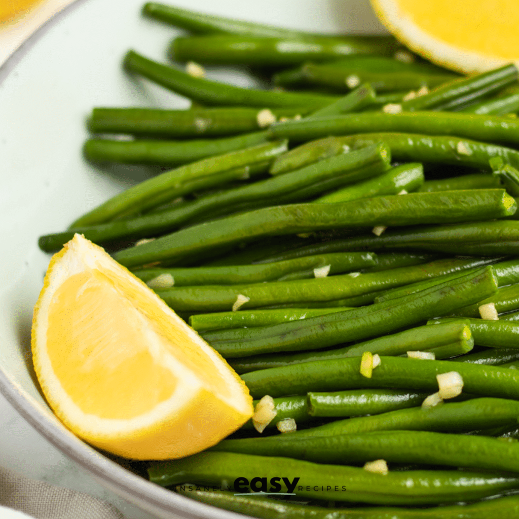 closeup of a bowl of whole green beans that have been sauteed with garlic. There are lemon wedges on the side of the bowl.