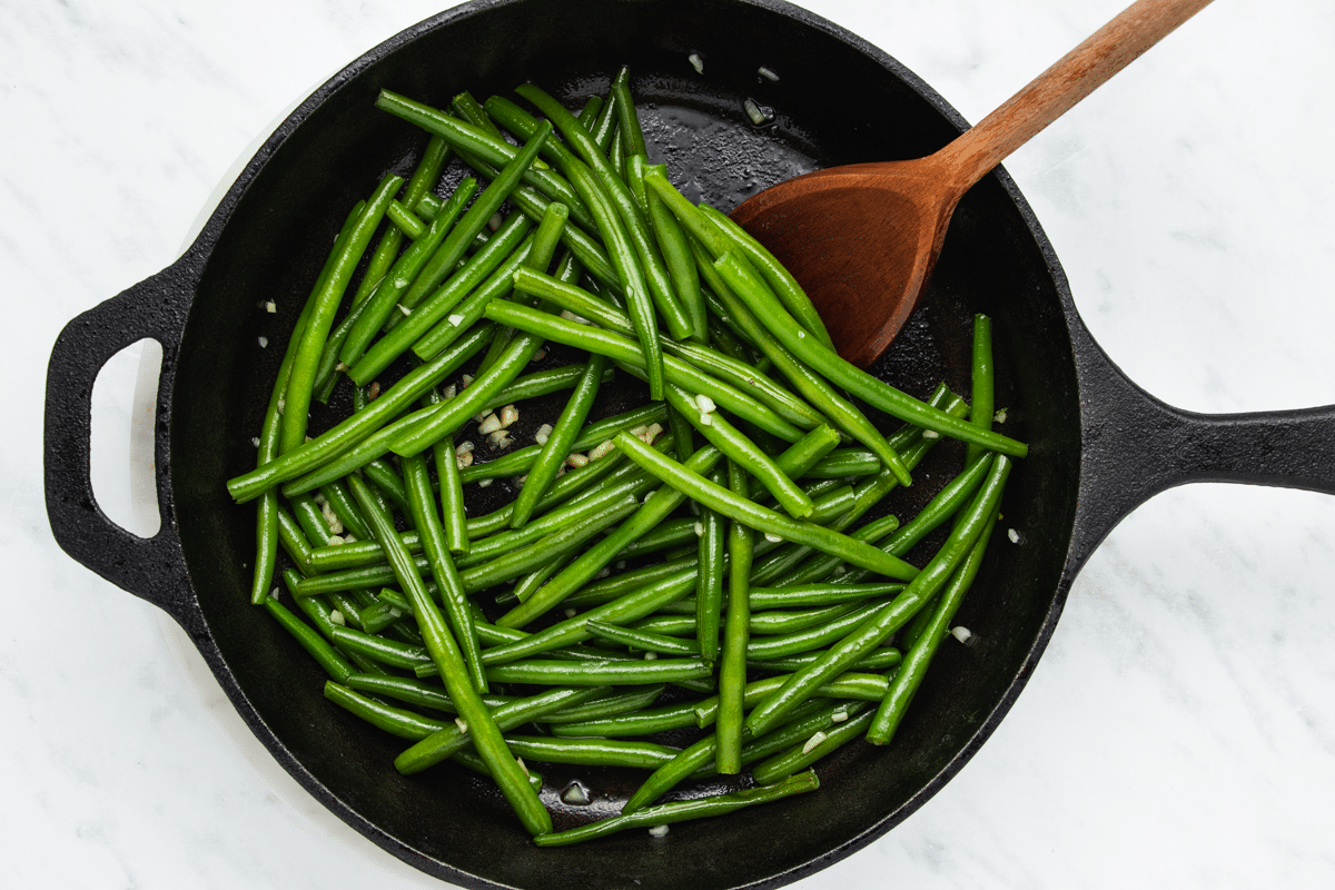 a cast iron skillet of green beans with garlic being stirred with a wooden spoon.