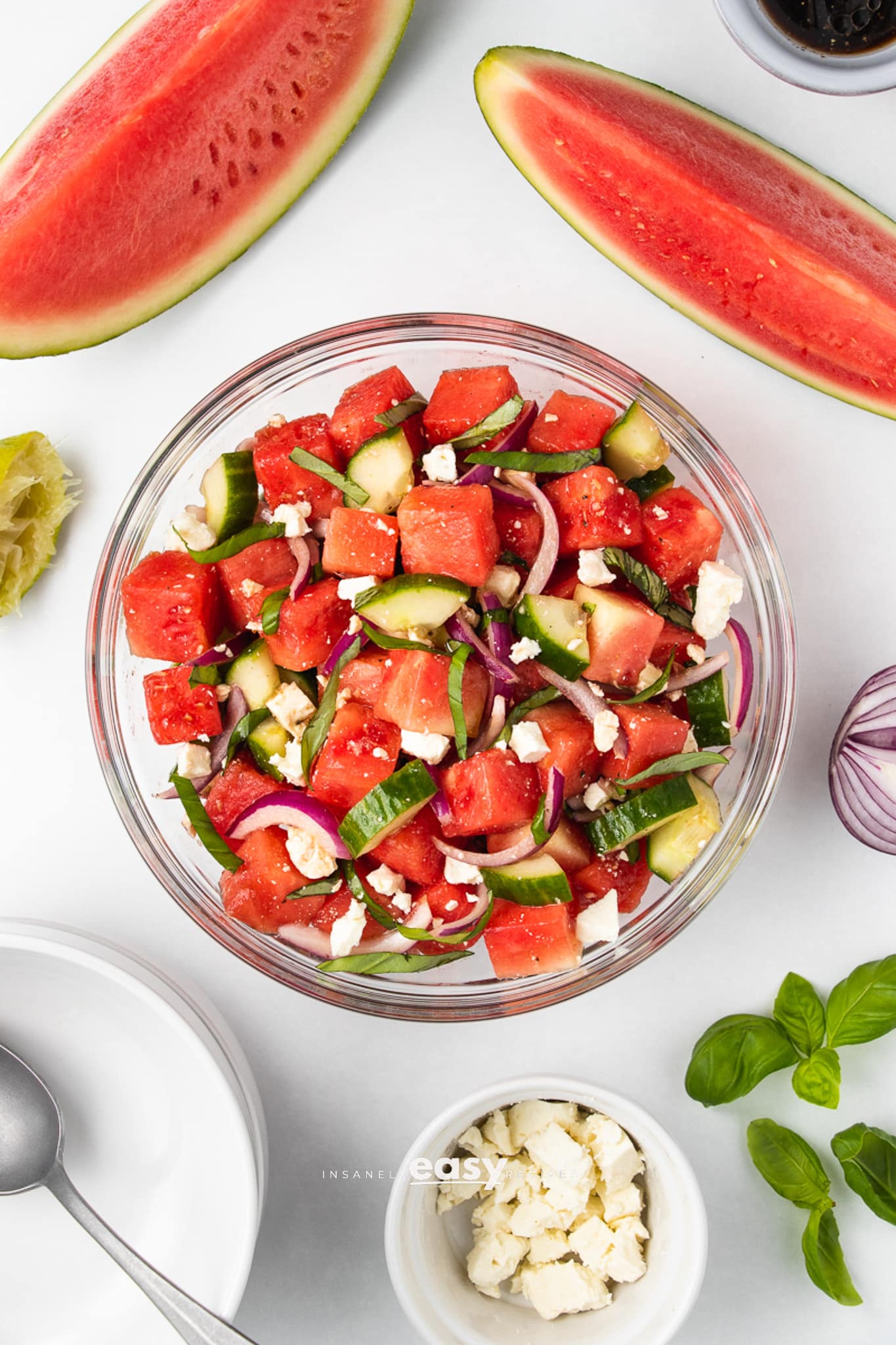 Top view photo of a large glass bowl filled with Watermelon Basil Salad. There are watermelon quarters at the top of the photo and basil and feta cheese  in bowls at the bottom of the photo. 